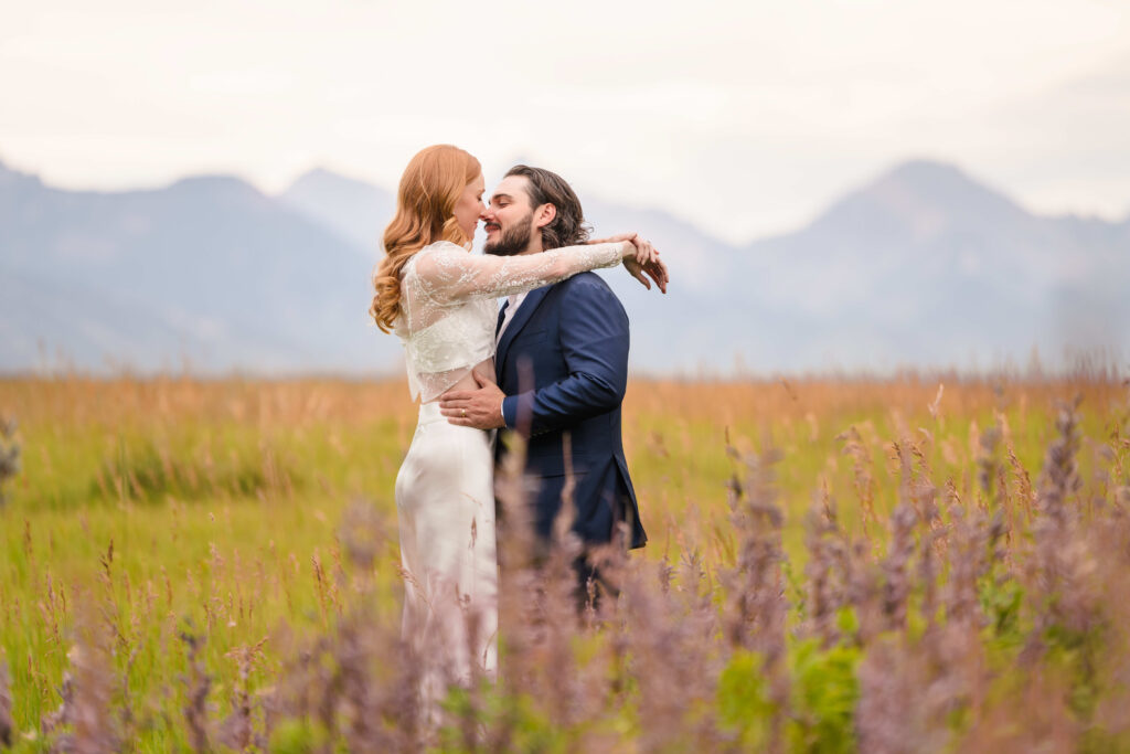A bride and groom stand close together in tall golden grass, pausing just before a kiss as distant mountain peaks frame the background.
