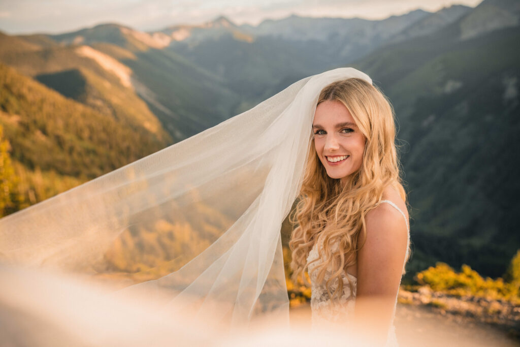 Bride standing in front of Colorado mountains as her veil blows in the wind and she smiles.