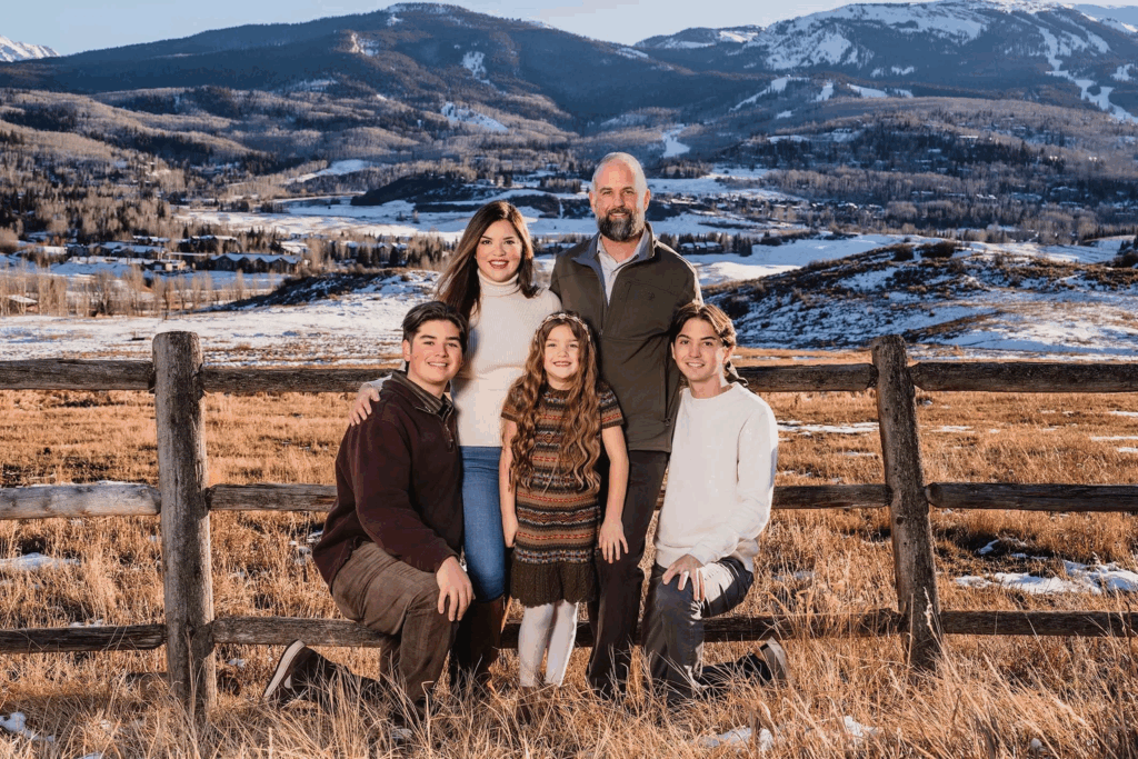 A family made up of a husband, wife, and 3 children pose in front of a fence with the snow-capped Aspen mountains behind them.
