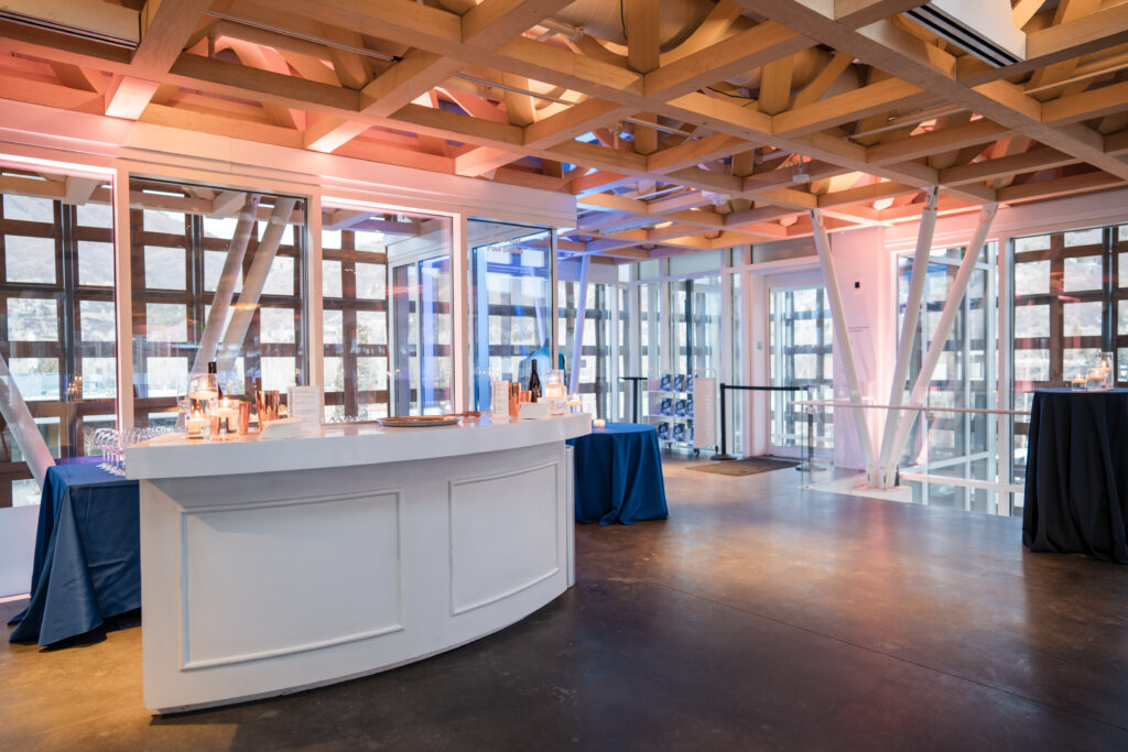 A sleek, white curved bar and blue cocktail tables set up for an evening reception at the Aspen Art Museum, showcasing professional corporate event photography. The shot highlights the venue's iconic wooden lattice ceiling, expansive glass walls, and warm ambient lighting.