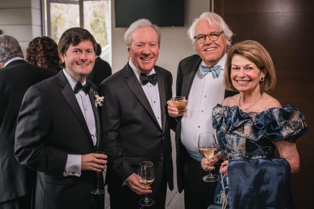 Elegant indoor event group portrait of four well-dressed guests in formal attire holding drinks and smiling at the camera, a candid and polished Photo Booth Alternative capturing natural interactions.